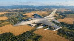 A fighter jet flies over a rural landscape with fields, trees, and scattered houses visible below.