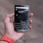 A person holds a BlackBerry Porsche Design smartphone with a physical keyboard and screen displaying the time.