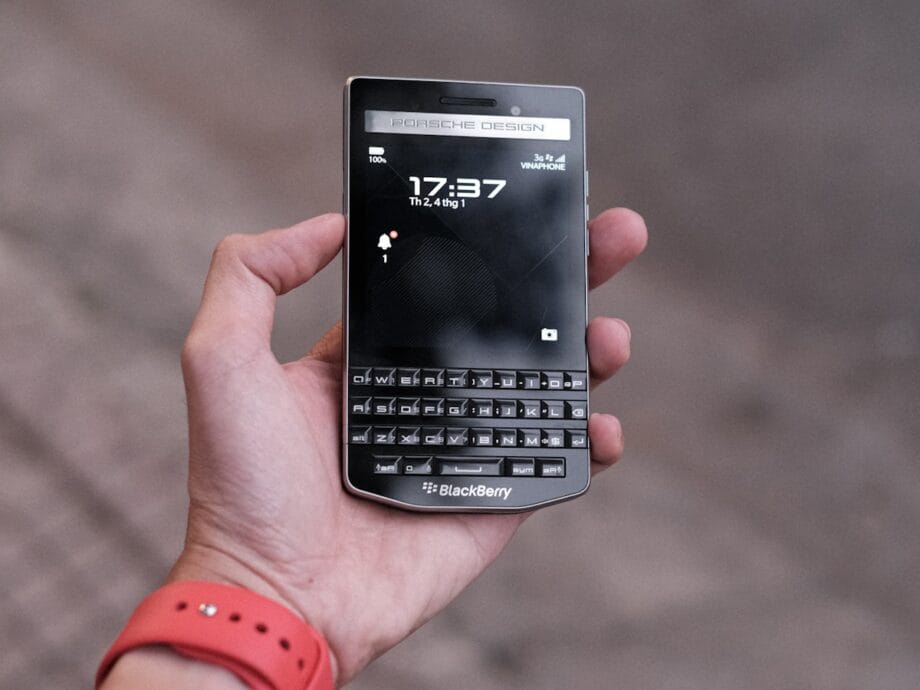 A person holds a BlackBerry Porsche Design smartphone with a physical keyboard and screen displaying the time.