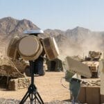 Military anti-drone radar and sensor equipment deployed in a desert base, with a flying drone and vehicles in the background.