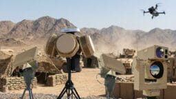 Military anti-drone radar and sensor equipment deployed in a desert base, with a flying drone and vehicles in the background.