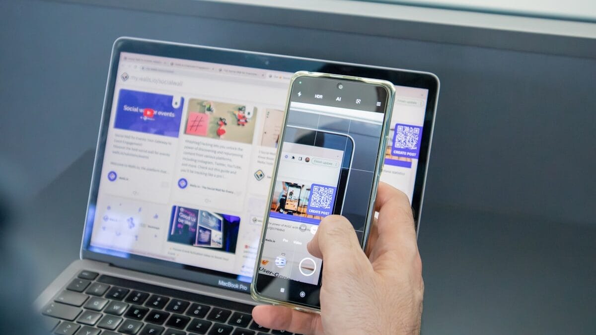 Person holding a smartphone to scan a QR code displayed on a laptop screen showing a website with social media content.