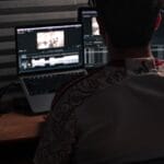 A person sits at a desk editing video footage on two laptop screens in a dimly lit room with grey soundproof panels.