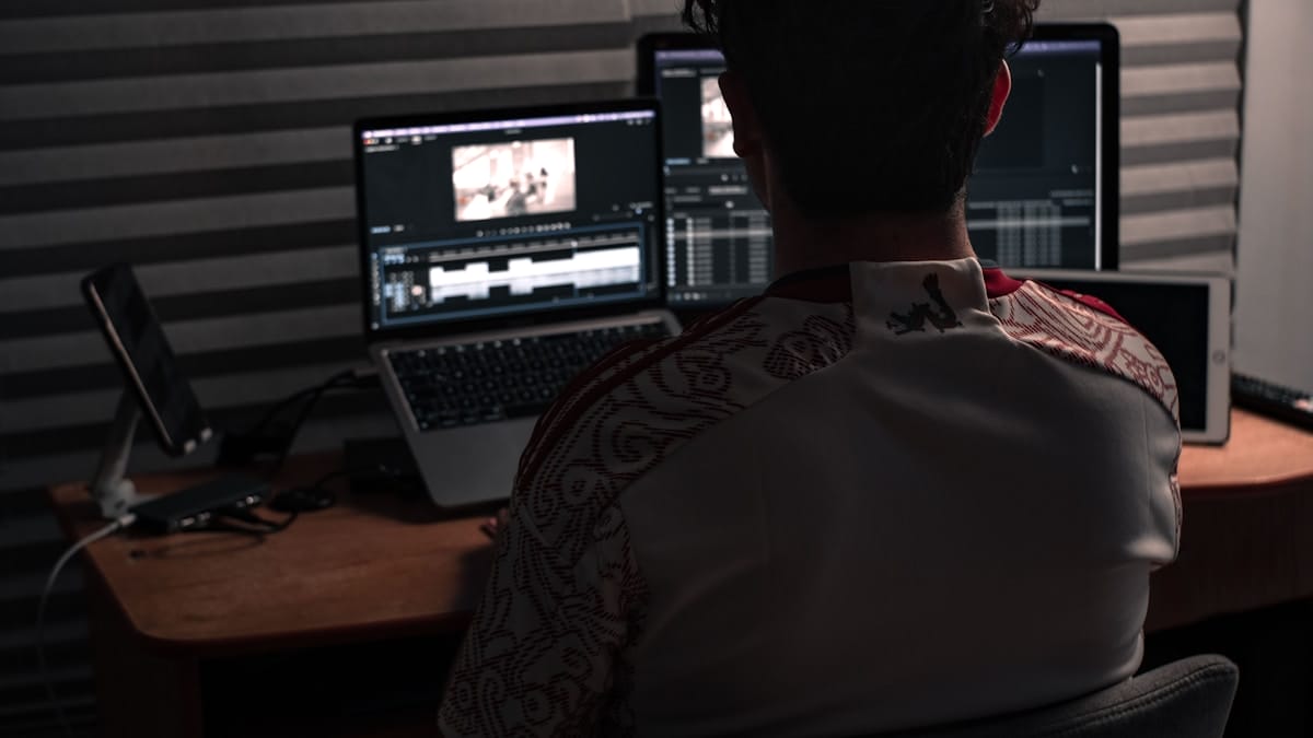 A person sits at a desk editing video footage on two laptop screens in a dimly lit room with grey soundproof panels.