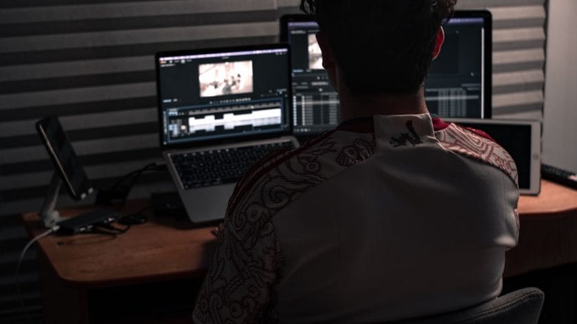 A person sits at a desk editing video footage on two laptop screens in a dimly lit room with grey soundproof panels.
