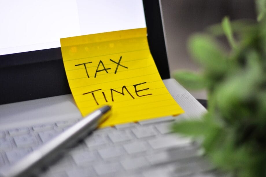 A sticky note with TAX TIME written on it is placed on a laptop keyboard next to a pen and a blurred plant.