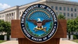 Podium with the seal of the U.S. Department of Defense in front of a government building and greenery.
