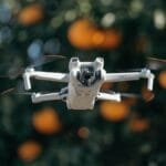 A small white drone with four propellers hovers outdoors in front of blurred orange and green foliage.