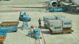 Airport ground crew moves metal cargo containers near an airplane on the tarmac, with equipment and carts nearby.