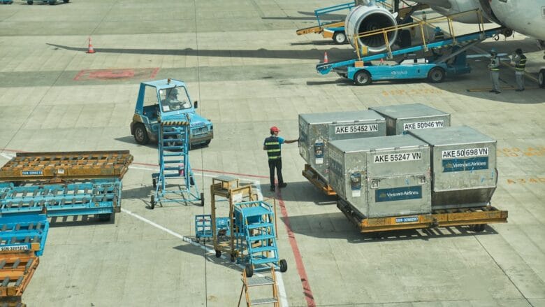 Airport ground crew moves metal cargo containers near an airplane on the tarmac, with equipment and carts nearby.