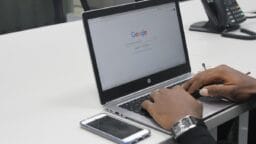 Person typing on a laptop with Google homepage on screen; smartphone and office phone on the white desk.