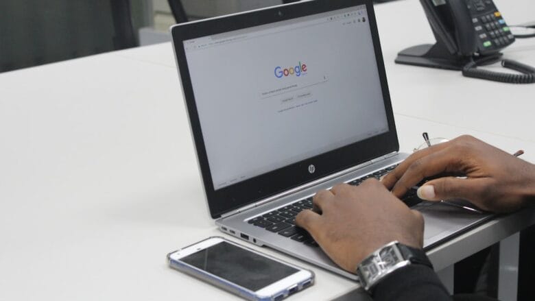 Person typing on a laptop with Google homepage on screen; smartphone and office phone on the white desk.
