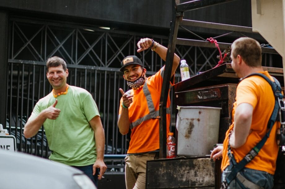 Three construction workers in safety gear smile and gesture toward the camera while standing next to equipment outdoors.