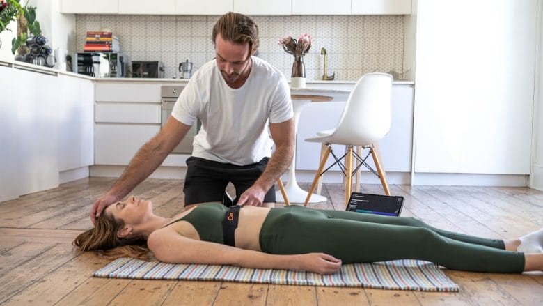 A man checks the pulse of a woman lying on a mat in a kitchen, wearing workout clothes and a fitness monitor.