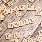 Scrabble tiles arranged to spell UNITY on a wooden surface, with other random letter tiles scattered around.