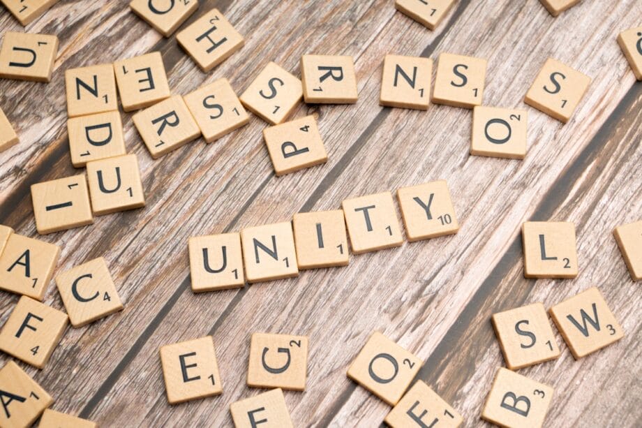 Scrabble tiles arranged to spell UNITY on a wooden surface, with other random letter tiles scattered around.