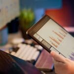 Person holding a tablet displaying bar and line graphs, with a desktop monitor showing charts in the background on a desk.