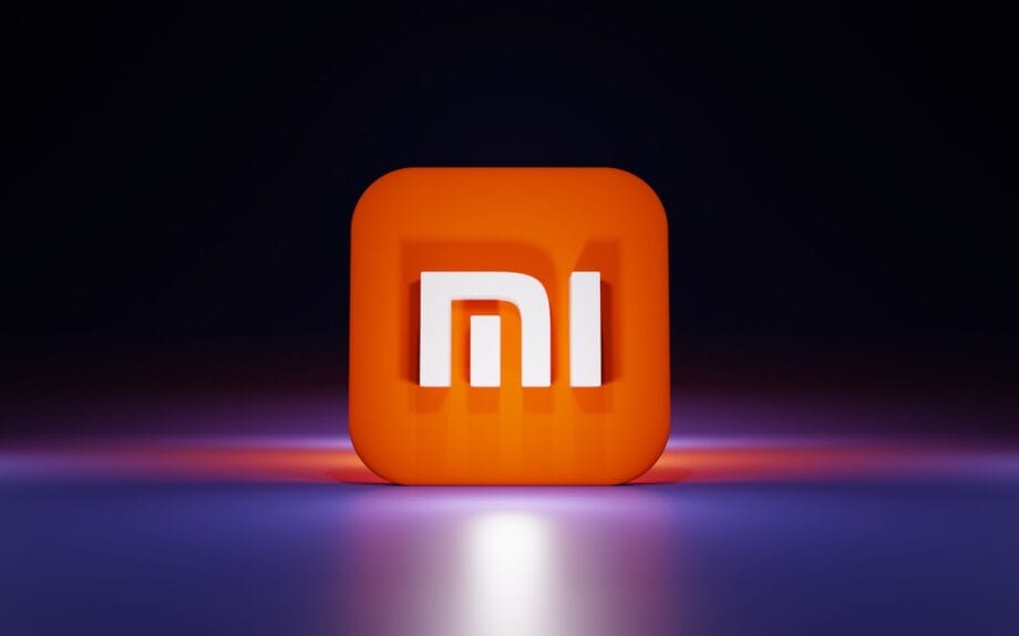 A 3D orange square with the white Xiaomi mi logo sits on a reflective surface against a dark background.