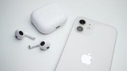 White wireless earbuds with charging case next to a white smartphone with the Apple logo on a white surface.