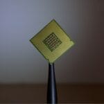 A computer processor chip held upright by black tweezers against a blurred background.