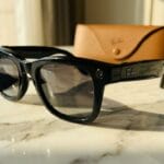 Black Ray-Ban sunglasses on a marble table with a brown Ray-Ban case and a cleaning cloth nearby.