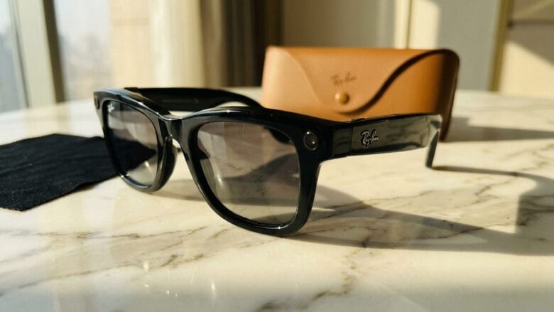 Black Ray-Ban sunglasses on a marble table with a brown Ray-Ban case and a cleaning cloth nearby.