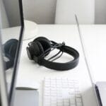 Black headphones rest on a white desk between a computer monitor, a wireless keyboard, and a laptop.