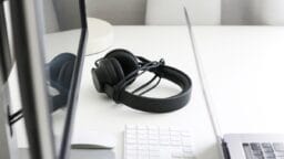 Black headphones rest on a white desk between a computer monitor, a wireless keyboard, and a laptop.