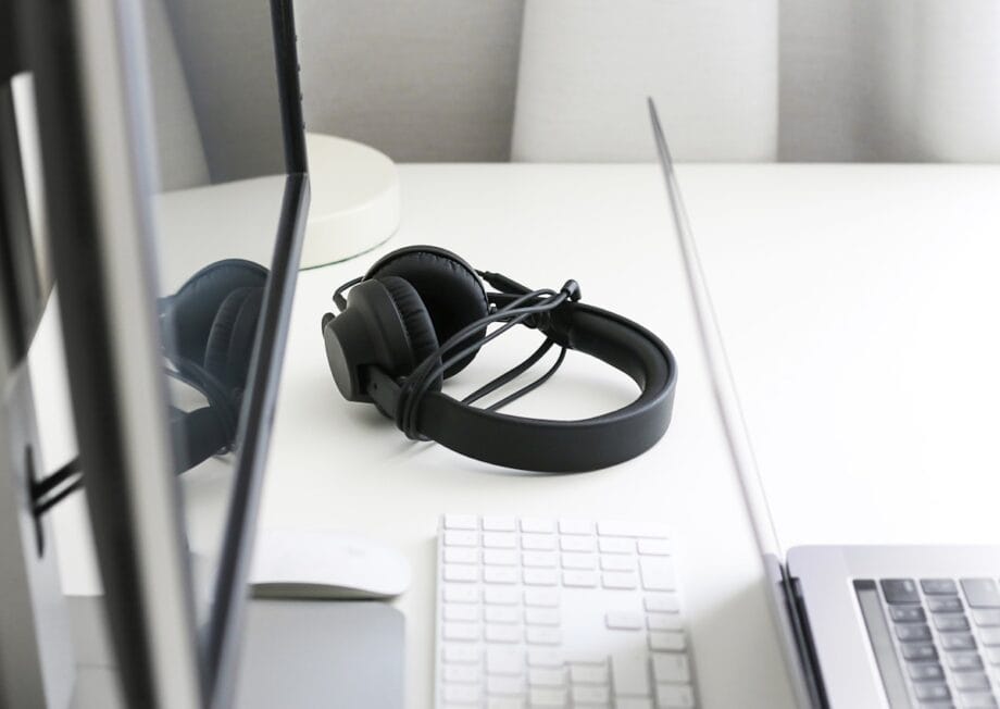 Black headphones rest on a white desk between a computer monitor, a wireless keyboard, and a laptop.