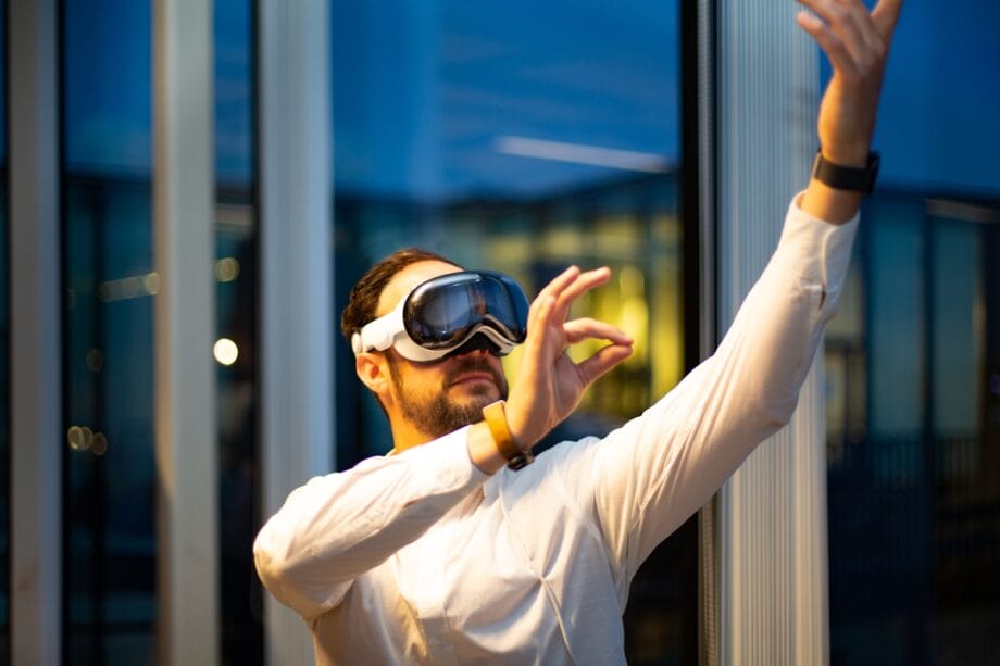 A man wearing a white shirt and VR headset raises one hand and gestures, standing indoors near large windows.