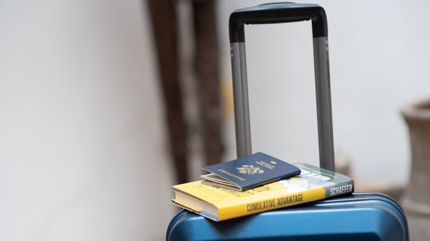 A blue suitcase with two books and a dark blue passport placed on top, against a neutral indoor background.