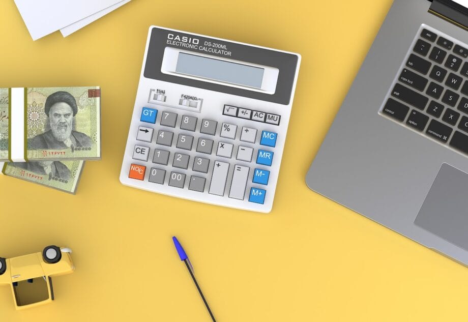 A calculator, laptop, Iranian banknotes, pen, and yellow toy car arranged on a yellow surface.