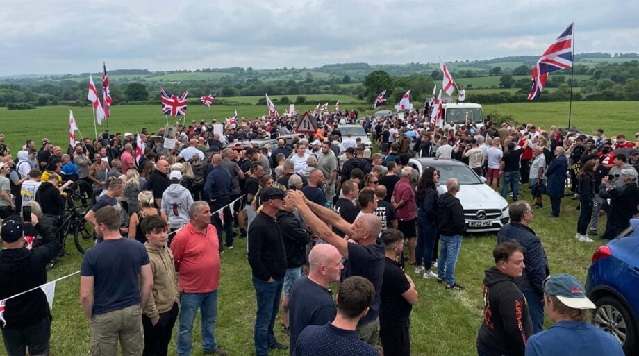 A large crowd gathers outdoors with numerous British flags and vehicles on a grassy field under a cloudy sky.