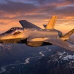 A fighter jet flies above snow-capped mountains and a winding river at sunset under a partly cloudy sky.