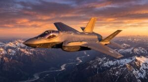 A fighter jet flies above snow-capped mountains and a winding river at sunset under a partly cloudy sky.