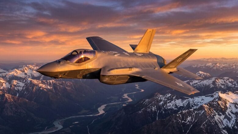 A fighter jet flies above snow-capped mountains and a winding river at sunset under a partly cloudy sky.