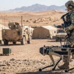 A man in military uniform holding a device in front of a desert landscape.