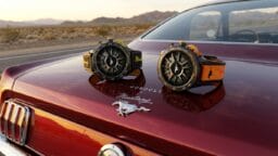 Two wristwatches are displayed on the trunk of a classic Ford Mustang parked in a desert landscape.