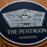 A large sign of the Pentagon building and text reading The Pentagon Washington mounted on a wooden wall beside a U.S. flag.