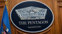 A large sign of the Pentagon building and text reading The Pentagon Washington mounted on a wooden wall beside a U.S. flag.