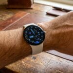 A person wearing a round-faced smartwatch with a white band is seated at a wooden desk.