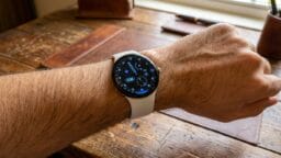 A person wearing a round-faced smartwatch with a white band is seated at a wooden desk.