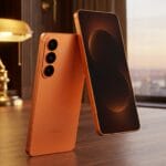 Two orange Samsung smartphones are displayed on a wooden desk in a warmly lit study with bookshelves and a window in the background.