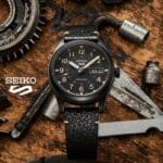A Seiko automatic wristwatch with a black dial and leather strap is displayed on a wooden surface amid metal gears and tools.