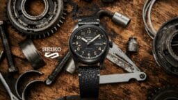 A Seiko automatic wristwatch with a black dial and leather strap is displayed on a wooden surface amid metal gears and tools.