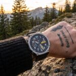A person wearing a black watch and sweater rests their hand on a rocky surface with mountains and pine trees in the background.