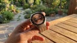 A hand holds a Huawei smartwatch displaying the time 18:32, with a garden and wooden table in the background.