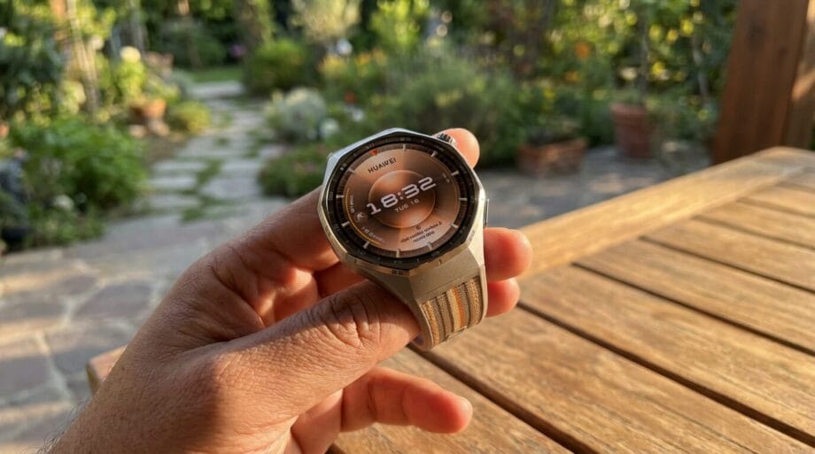 A hand holds a Huawei smartwatch displaying the time 18:32, with a garden and wooden table in the background.