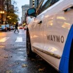 A Waymo self-driving car is parked on a city street near a crosswalk at dusk with pedestrians and buildings in the background.