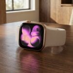 A smart watch with a pink abstract display rests on a wooden table in a sunlit room.